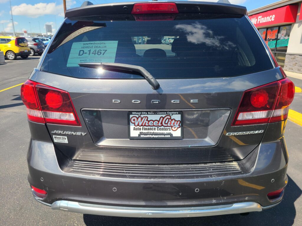 Rear view of a dark gray Dodge Journey Crossroad SUV with a Wheel City Auto Finance Centers license plate frame visible, parked in a dealership lot with other cars and a red 'We Finance' sign in the background.