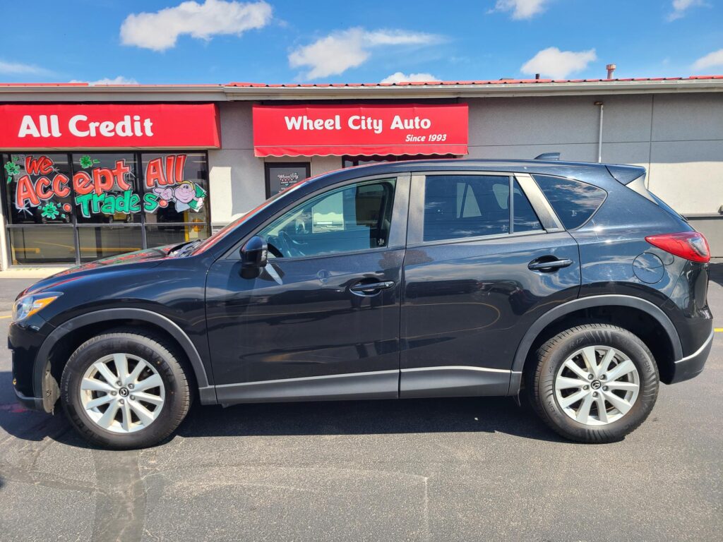 Front view of a car dealership with red signs reading 'Wheel City Auto' and 'All Credit'; a dark SUV is parked in front of the store.