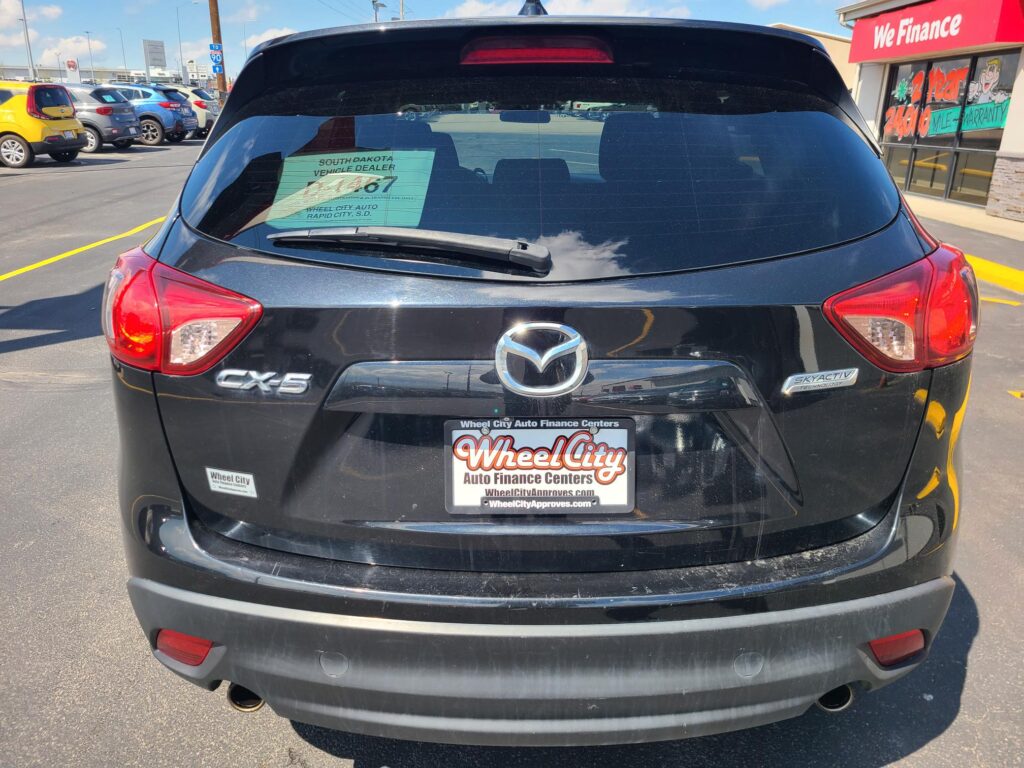 Rear view of a black Mazda CX-5 at a car lot with a green dealership window sticker on the rear windshield and Wheel City Auto Finance Centers license plate frame visible.