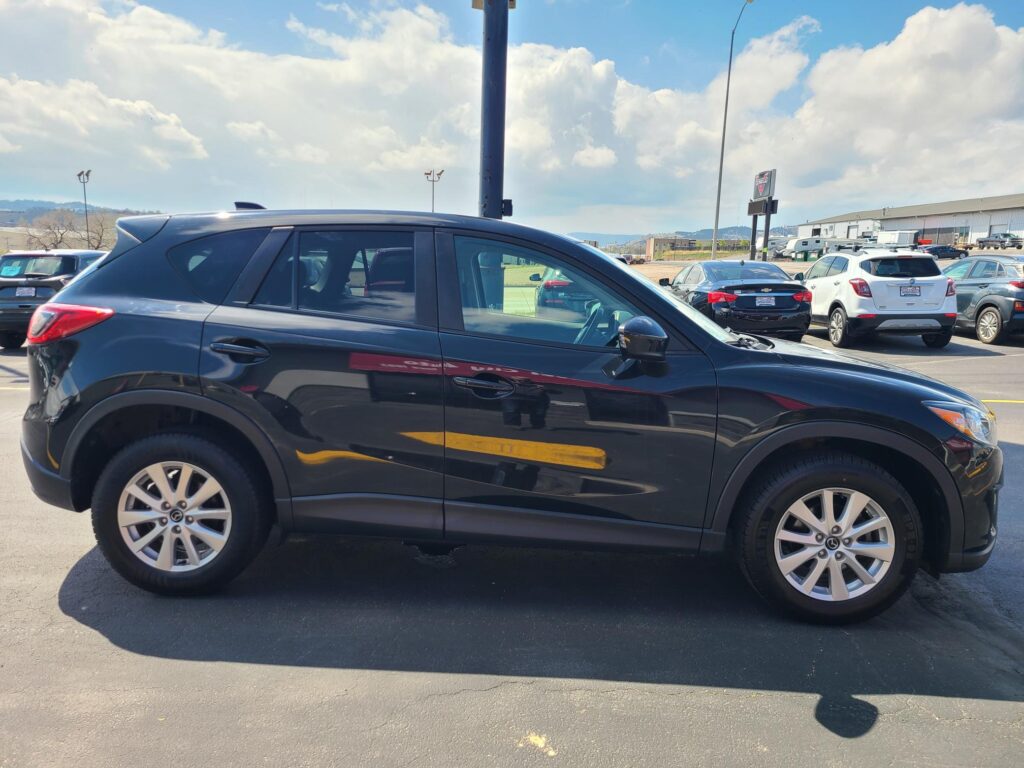 Close-up side view of a dark SUV parked in a parking lot, with other cars and distant industrial buildings under a partly cloudy sky.