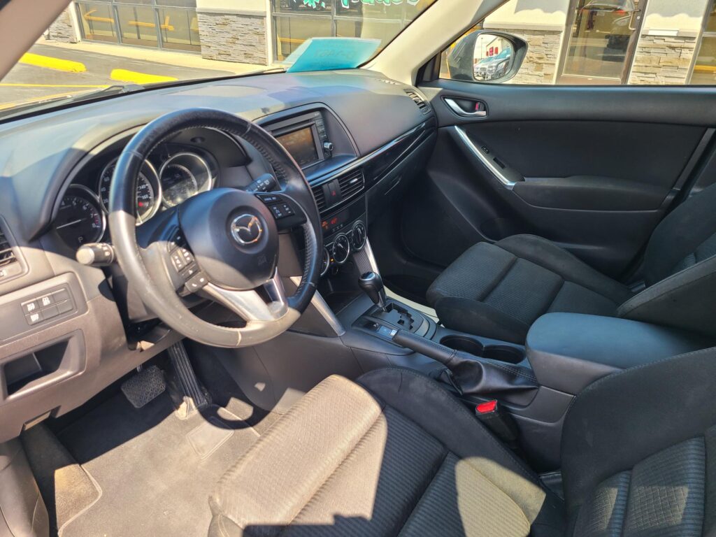 Interior of a Mazda car from the driver's seat showing steering wheel, dashboard, and center console with gear shifter and touchscreen display.