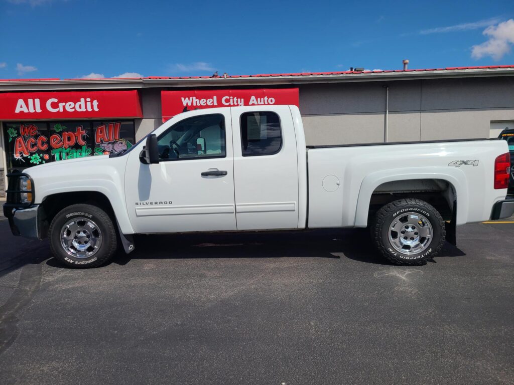White Chevrolet Silverado pickup parked in front of a storefront with red signs reading 'All Credit' and 'Wheel City Auto' under a blue sky; window graphic says 'We Accept Trades'.