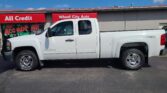 White Chevrolet Silverado pickup parked in front of a storefront with red signs reading 'All Credit' and 'Wheel City Auto' under a blue sky; window graphic says 'We Accept Trades'.