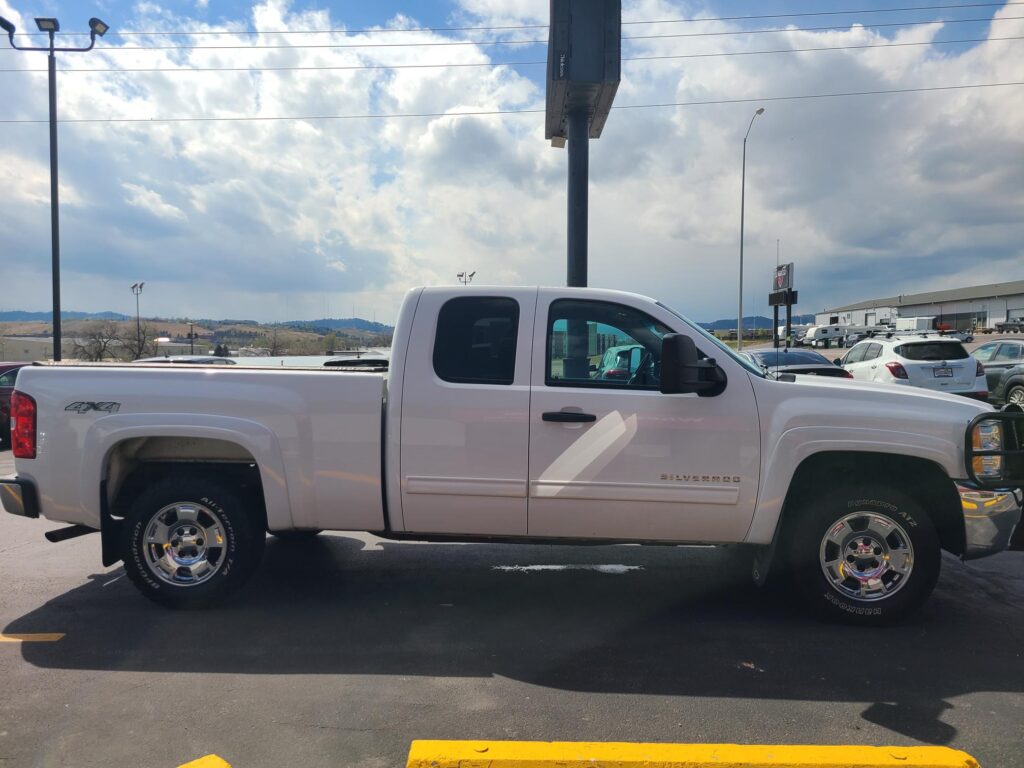 White Chevrolet Silverado pickup parked in a lot with yellow parking blocks and a cloudy blue sky above.