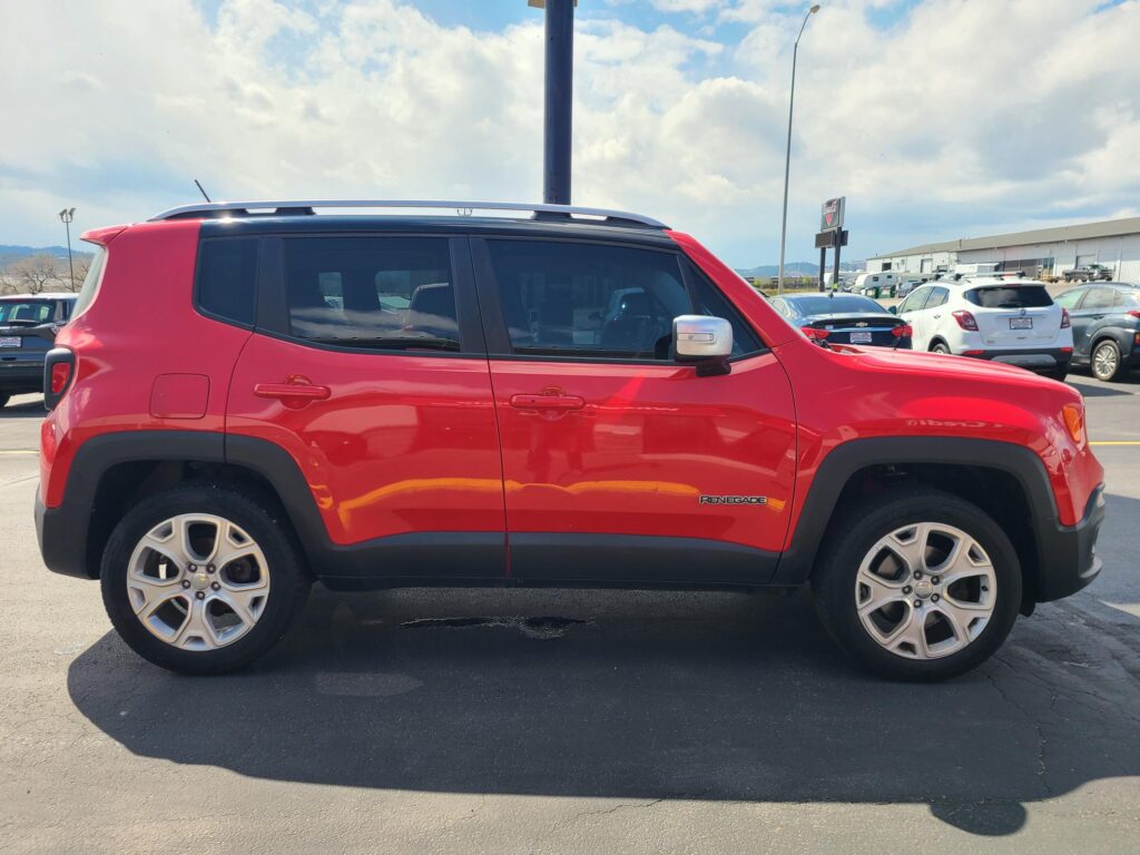 Red Jeep Renegade parked in a lot, side view showing doors, wheels, and roof rails with other cars and a dealership in the background.