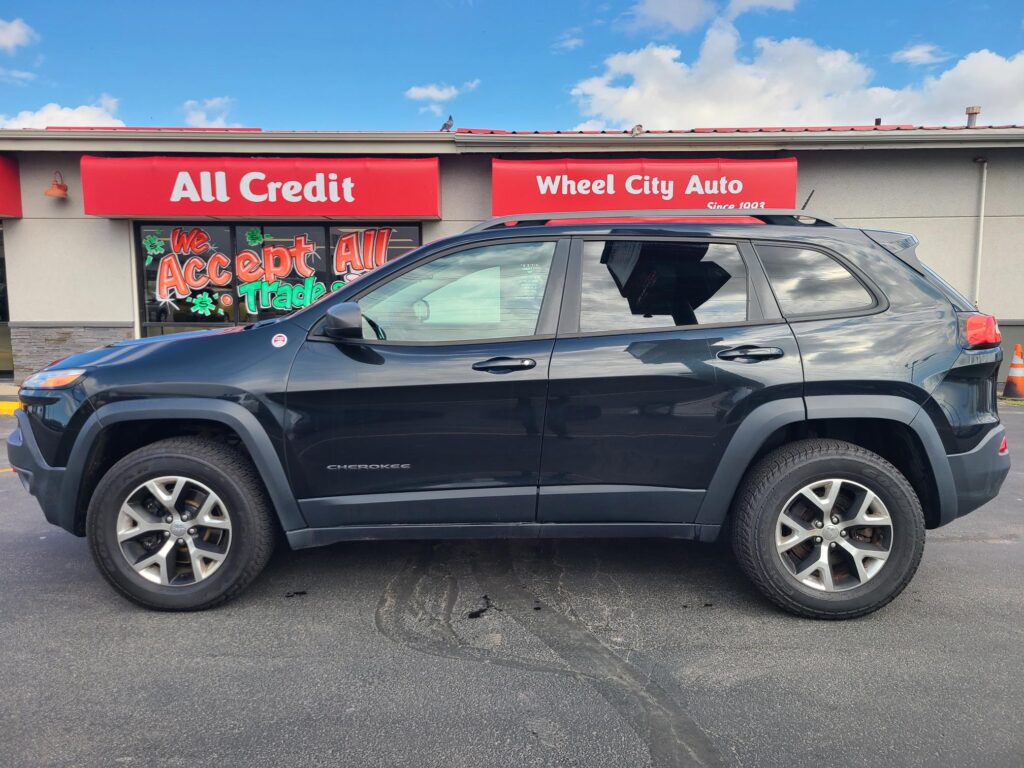 Blue SUV parked in front of a red-sign auto dealership with 'All Credit' and 'Wheel City Auto' storefront signs.