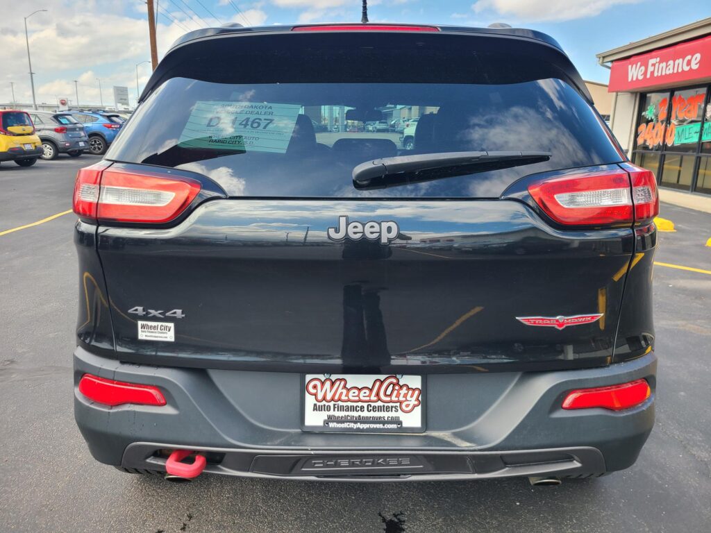 Rear view of a dark Jeep Cherokee 4x4 in a parking lot; visible dealer plate frame and tow hook, with a 'We Finance' storefront in the background.