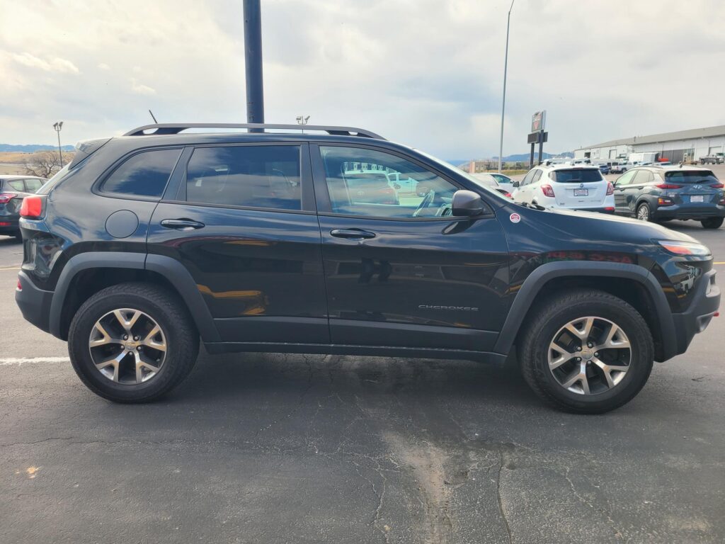 Black Jeep Cherokee SUV parked in a parking lot with other cars and an industrial building in the background.