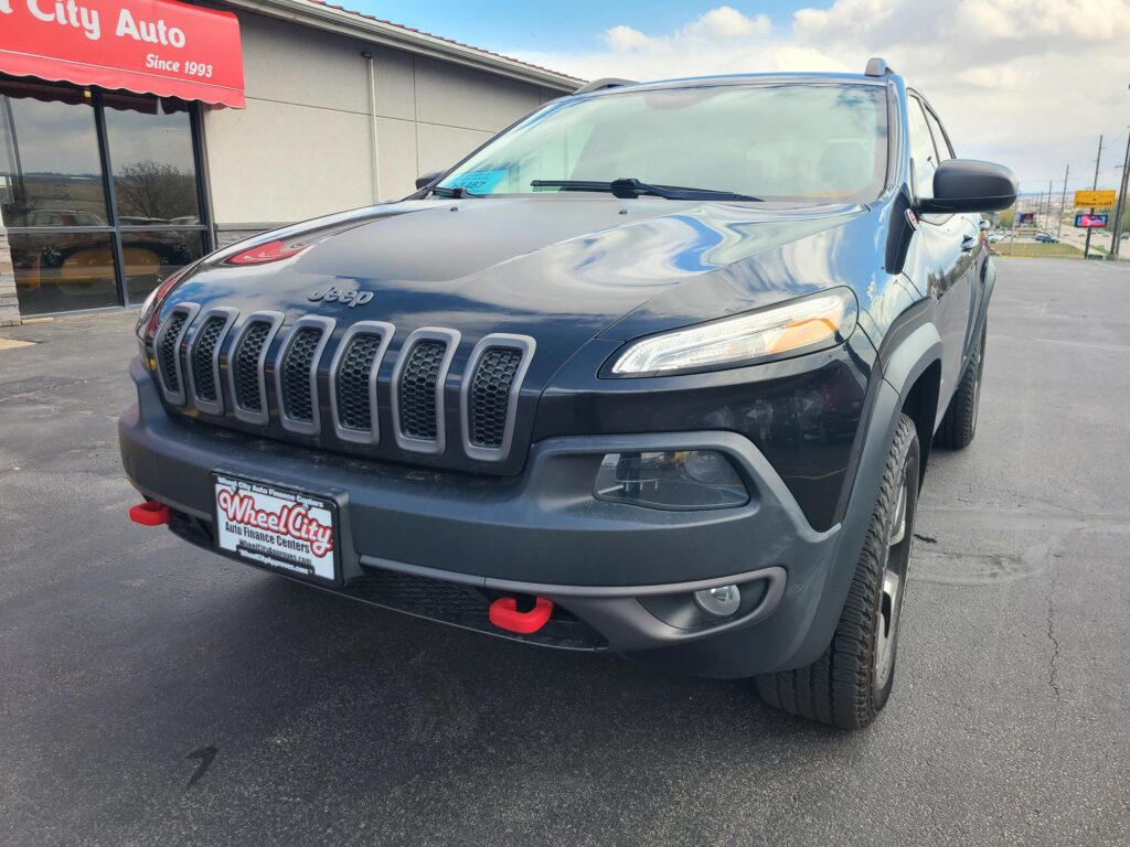 Front-quarter view of a dark gray Jeep SUV parked outside a Wheel City Auto dealership with a red canopy sign says 'Since 1993'