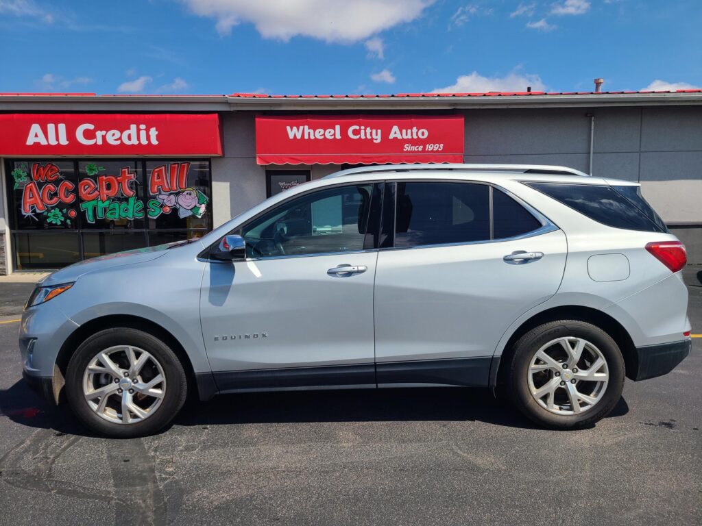 Silver SUV parked in front of a red storefront sign reading 'All Credit Wheel City Auto' under a blue sky.