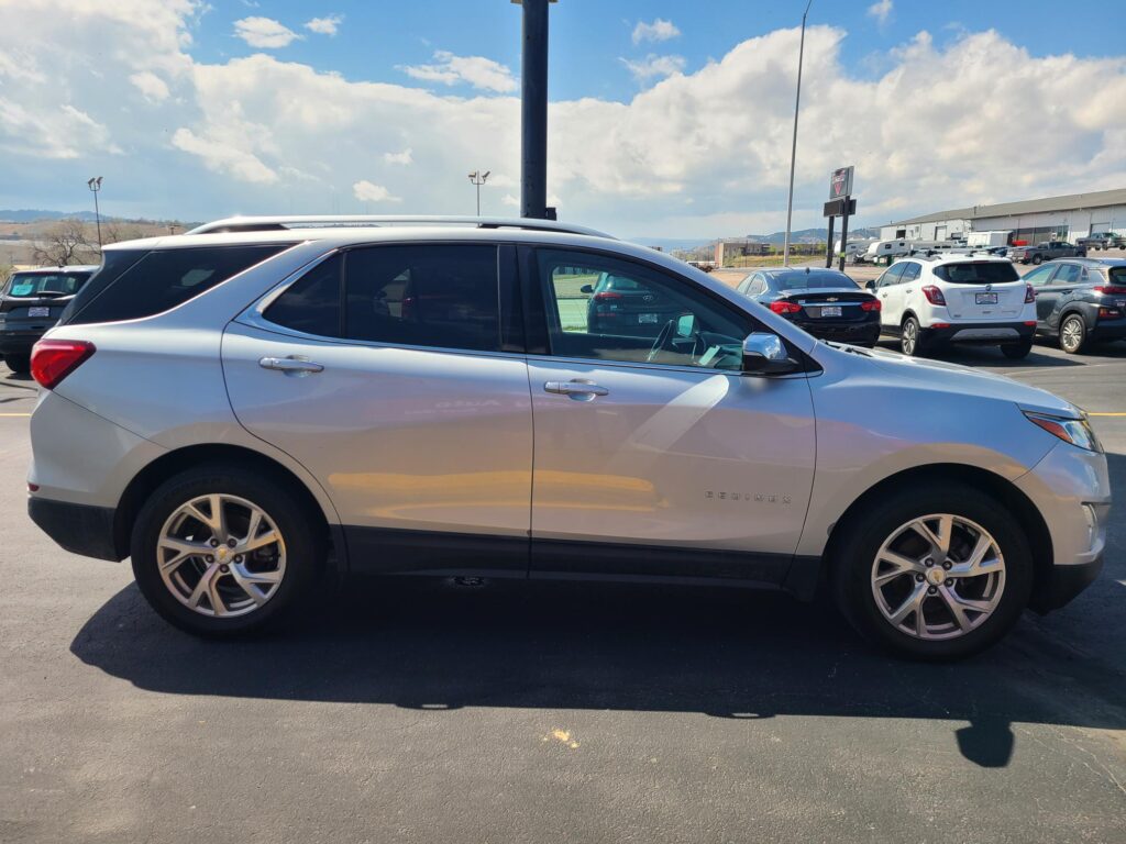 Silver Infiniti SUV (QX60) in a parking lot, photographed from the driver’s side with other cars and a building in the background under a partly cloudy sky.