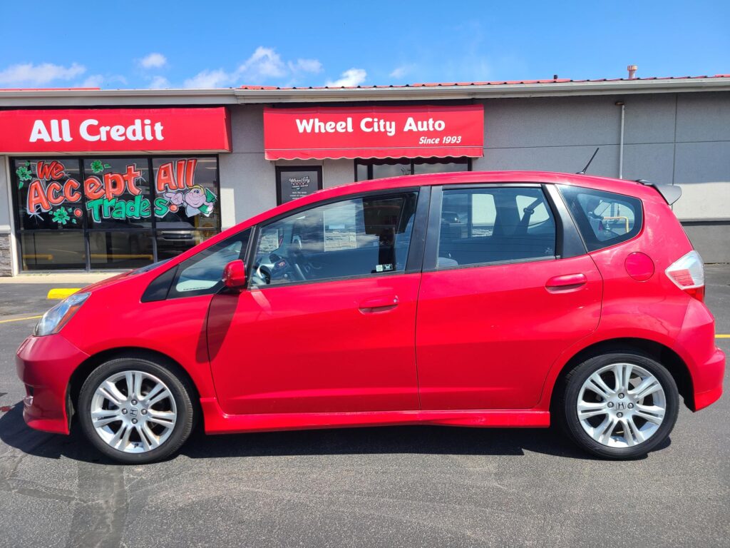 Red Honda hatchback parked in front of a storefront with a bold red 'All Credit' sign and a colorful mural reading 'We Accept All Trades'.