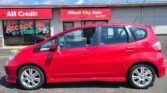 Red Honda hatchback parked in front of a storefront with a bold red 'All Credit' sign and a colorful mural reading 'We Accept All Trades'.
