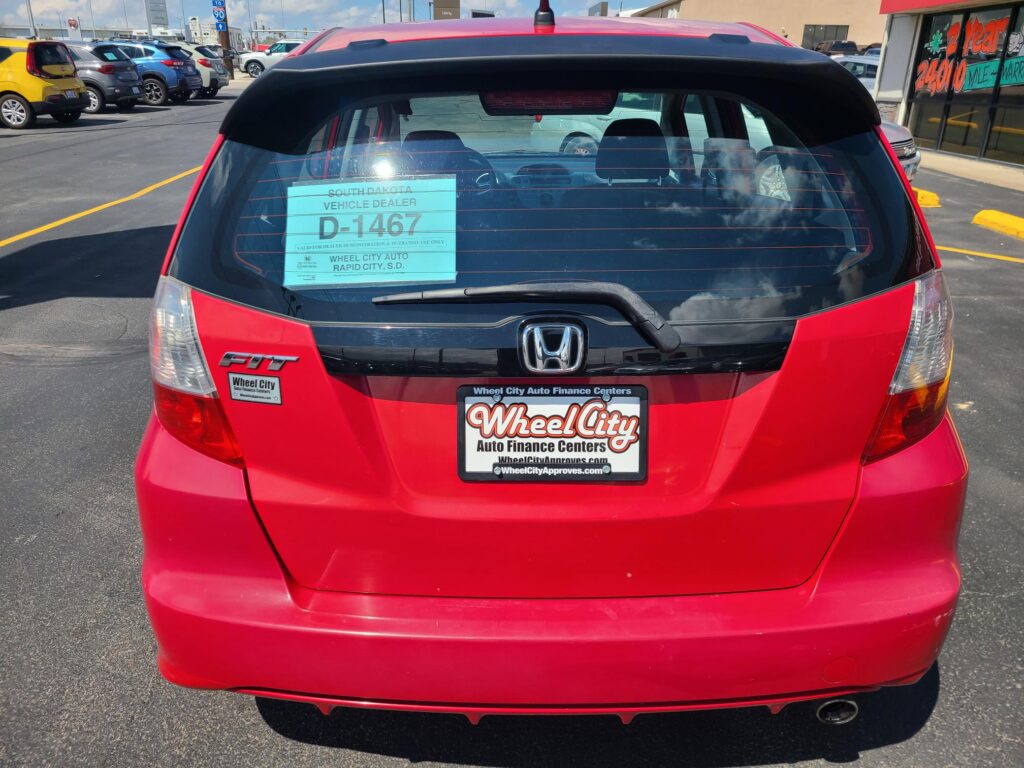 Rear view of a red Honda Fit at a car dealership, with a turquoise D-1467 dealer sticker visible on the rear window.