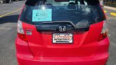 Rear view of a red Honda Fit at a car dealership, with a turquoise D-1467 dealer sticker visible on the rear window.