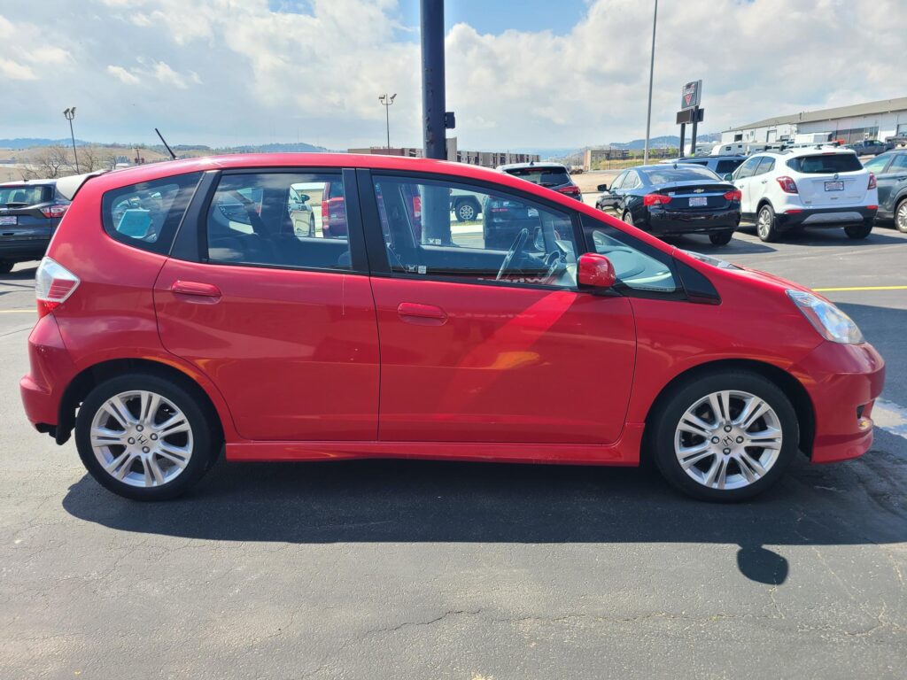 Red hatchback parked in a car dealership lot with other vehicles in the background on a sunny day
