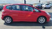 Red hatchback parked in a car dealership lot with other vehicles in the background on a sunny day