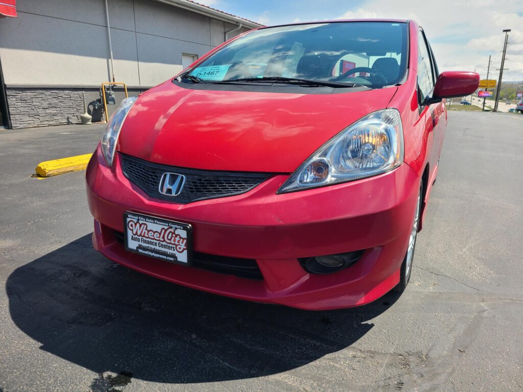 Front three-quarter view of a red Honda car, showing the headlight, grille, and windshield with a dealer sticker visible on the glass