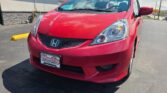 Front three-quarter view of a red Honda car, showing the headlight, grille, and windshield with a dealer sticker visible on the glass