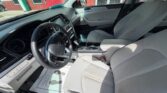 Interior of a gray sedan showing the driver's seat, steering wheel, dashboard, and center console with cupholders; front passenger seat and a building visible through the windows.
