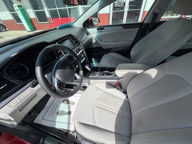 Interior of a gray sedan showing the driver's seat, steering wheel, dashboard, and center console with cupholders; front passenger seat and a building visible through the windows.