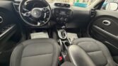 Front interior of a Kia car, view from the back seat: black cloth seats, steering wheel with Kia emblem, center console and infotainment screen visible.