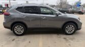 Side view of a gray compact SUV parked in a car lot; shows four doors, alloy wheels, and roof rails against a backdrop of other vehicles and dealership buildings.