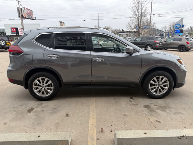 Side view of a gray compact SUV parked in a car lot; shows four doors, alloy wheels, and roof rails against a backdrop of other vehicles and dealership buildings.