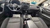 View of the car's front cabin: steering wheel on the left, center infotainment panel, gear shifter, and dark cloth seats viewed from the back seat of a Nissan car