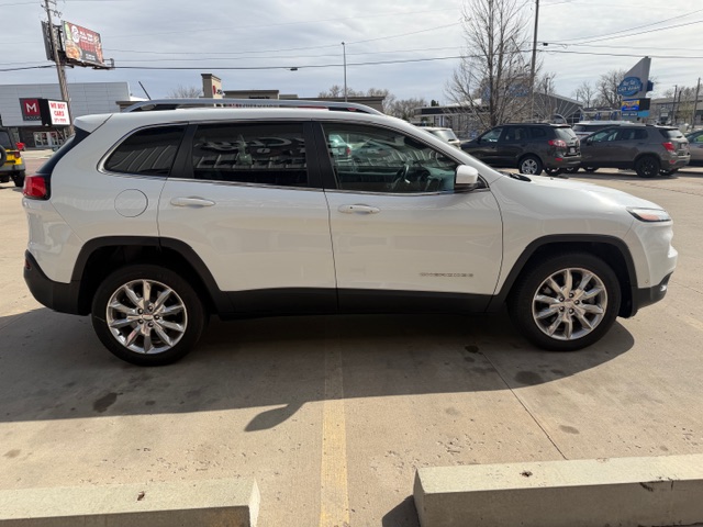 Side profile of a white mid-size SUV with roof rails and chrome wheels parked in a lot, other vehicles visible in the background.
