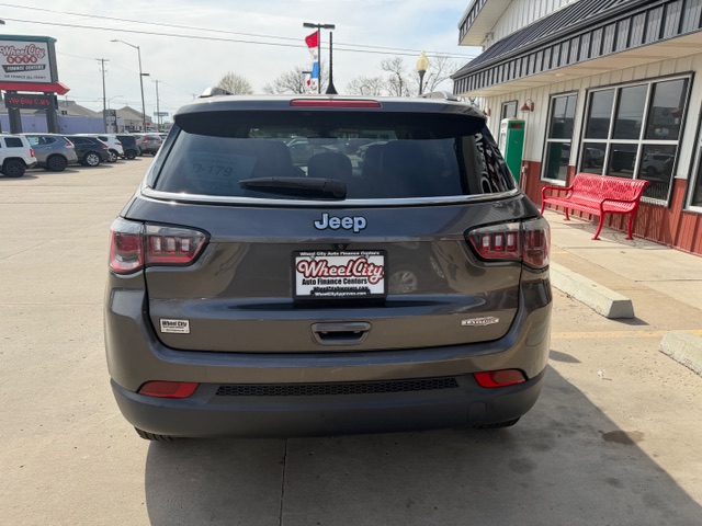Rear view of a gray Jeep SUV in a car dealership lot, with a Wheel City Auto Finance license plate frame and red bench outside a storefront to the right.