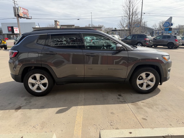 Side view of a gray Jeep Cherokee SUV parked in a parking lot at a dealership or mall area, showing the vehicle’s length and wheels.