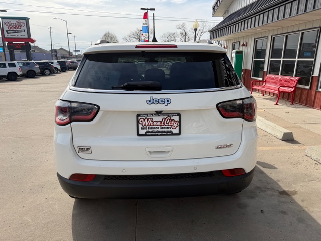 Rear view of a white Jeep SUV in a dealership lot; Wheel City license plate frame, red bench outside the storefront on the right.