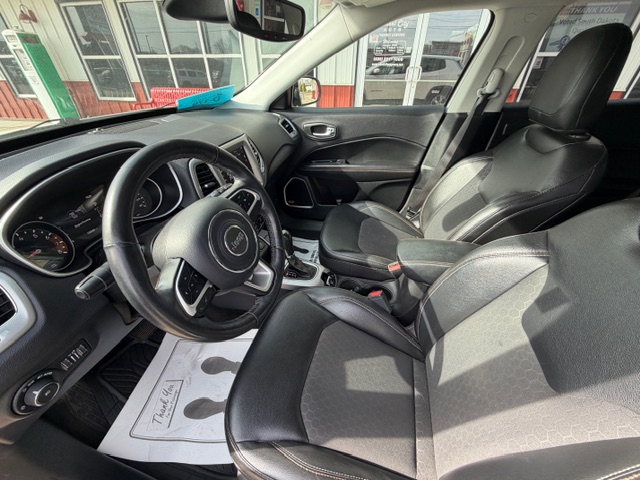 Front interior of a black Jeep SUV: steering wheel, dashboard gauges, two leather front seats, center console, and dealership paper on the floor mat.