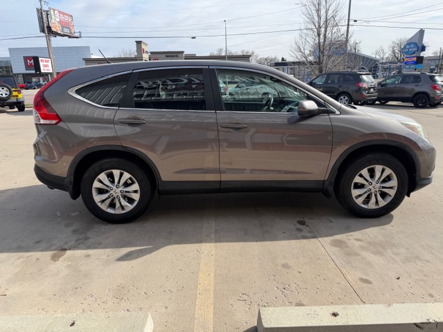 Side view of a brown-gray mid-size SUV parked in a dealership lot, with other cars and signs in the background.