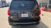 Rear view of a black Dodge Grand Caravan parked in a dealership lot, with a Wheel City license plate frame and a building with red benches to the right.