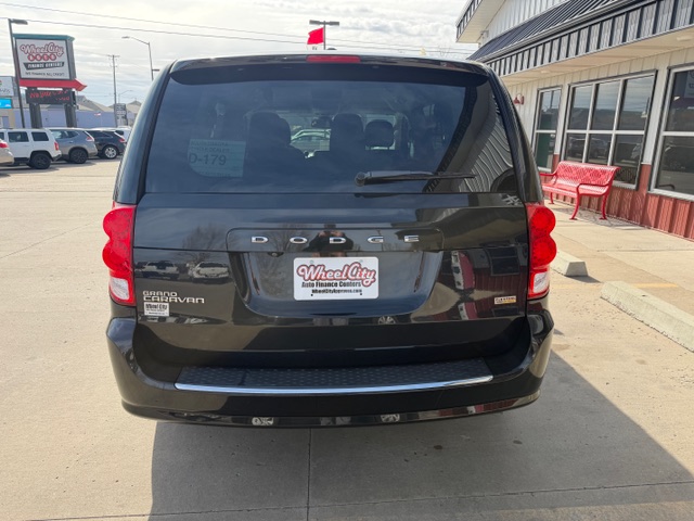 Rear view of a black Dodge Grand Caravan parked in a dealership lot, with a Wheel City license plate frame and a building with red benches to the right.