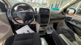 Front interior of a Dodge minivan: black cloth seats, steering wheel with logo, center console touchscreen, gear shifter area, and view of a dealership lot outside the windows.