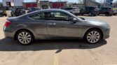 Gray two-door coupe parked in a sunny parking lot, with other cars and storefronts visible in the background.