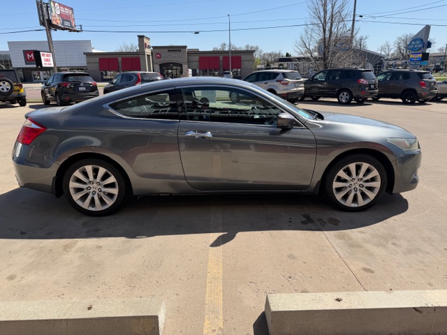 Gray two-door coupe parked in a sunny parking lot, with other cars and storefronts visible in the background.