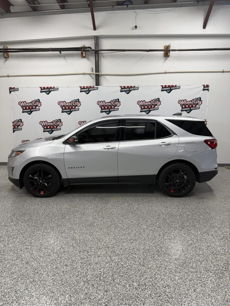 Silver SUV (Equinox) parked indoors in front of a banner with Wheel City Auto Finance Centers logos on a white wall backdrop.