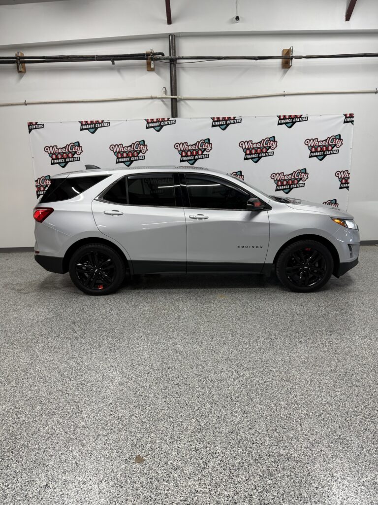 Silver Chevrolet Equinox SUV parked indoors in a showroom with a Wheel City Auto Finance Centers backdrop banner behind it.