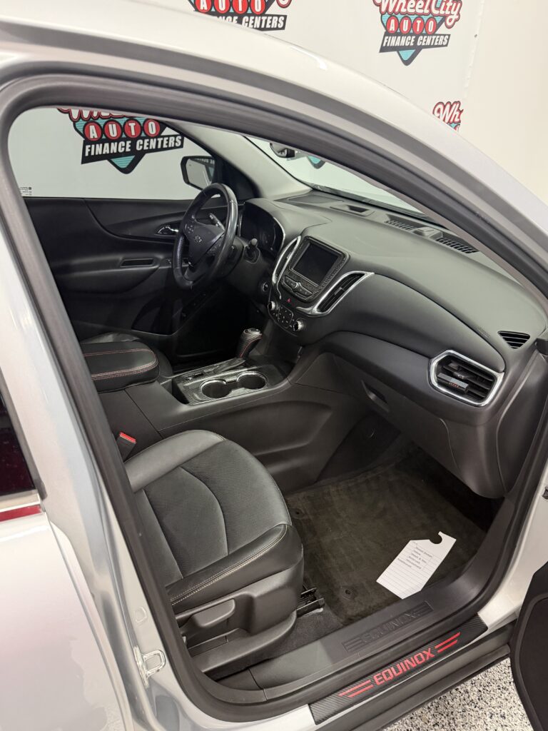 Interior of a Chevrolet Equinox driver’s area: black seats, center console, steering wheel, and dashboard with a touchscreen display visible from the open front door.