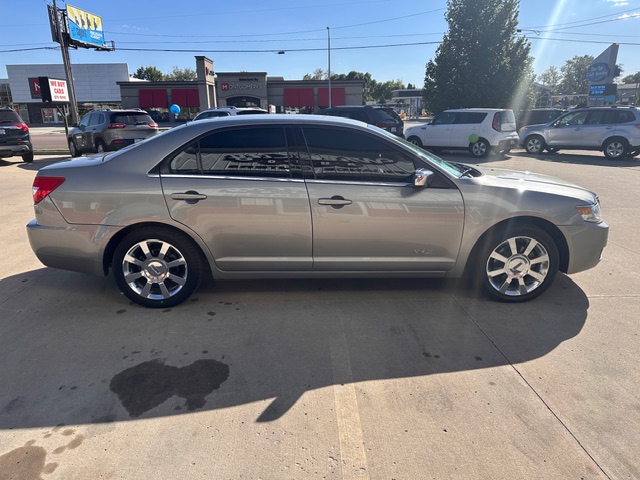 Side view of a silver four-door sedan parked in a car dealership lot with other cars in the background.