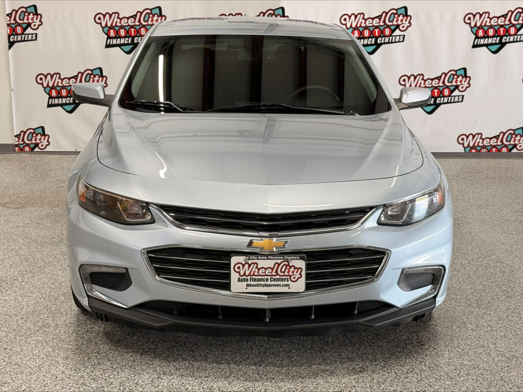 Front view of a silver Chevrolet sedan in a showroom with Wheel City Auto Finance Centers backdrop on the wall behind it.