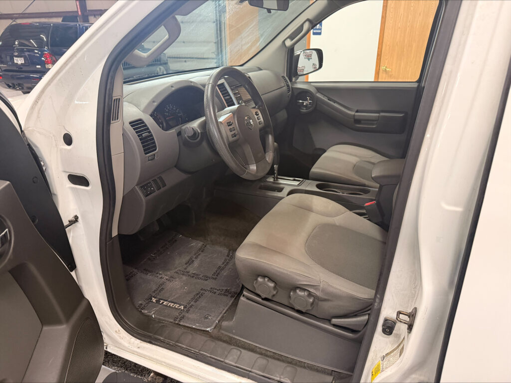 Interior of a white pickup truck; driver’s seat, steering wheel, center console with gear shifter, and passenger seat visible through the open door.