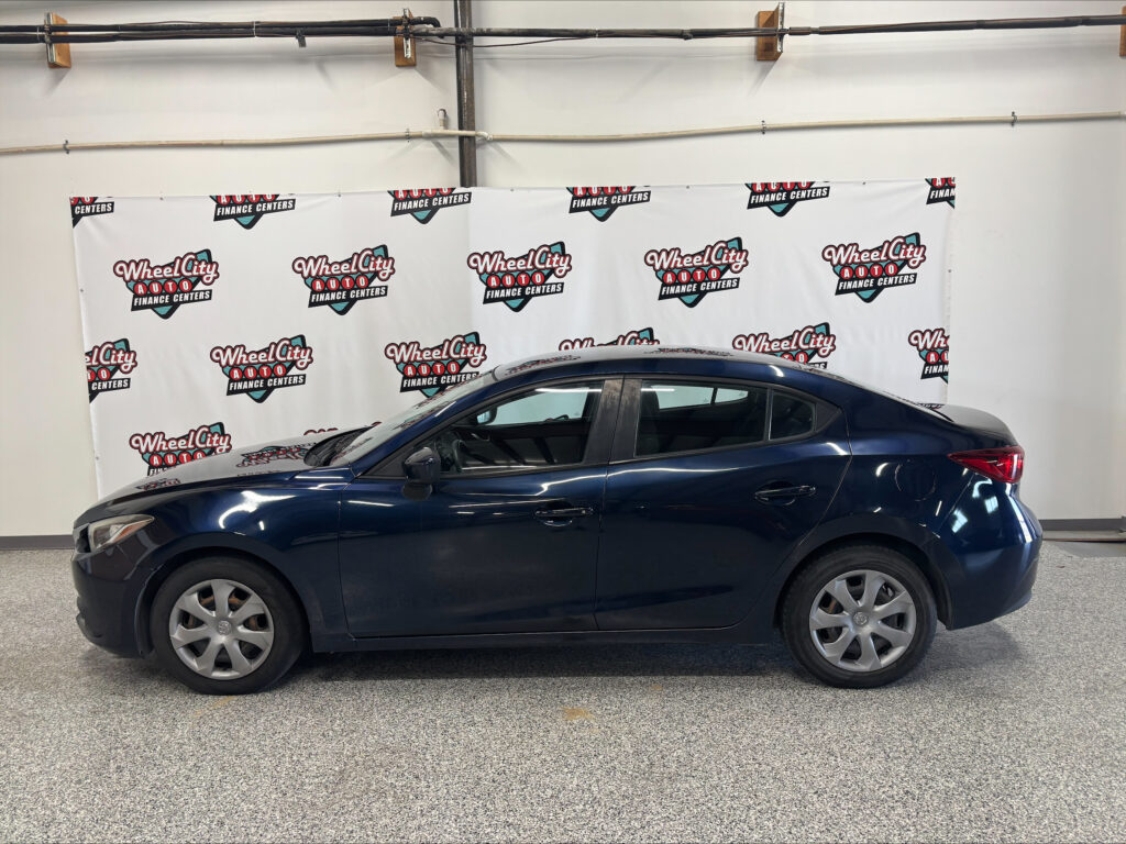 Side view of a dark blue sedan parked indoors in a showroom with a white wall and Wheel City Auto Finance Centers banners behind it.