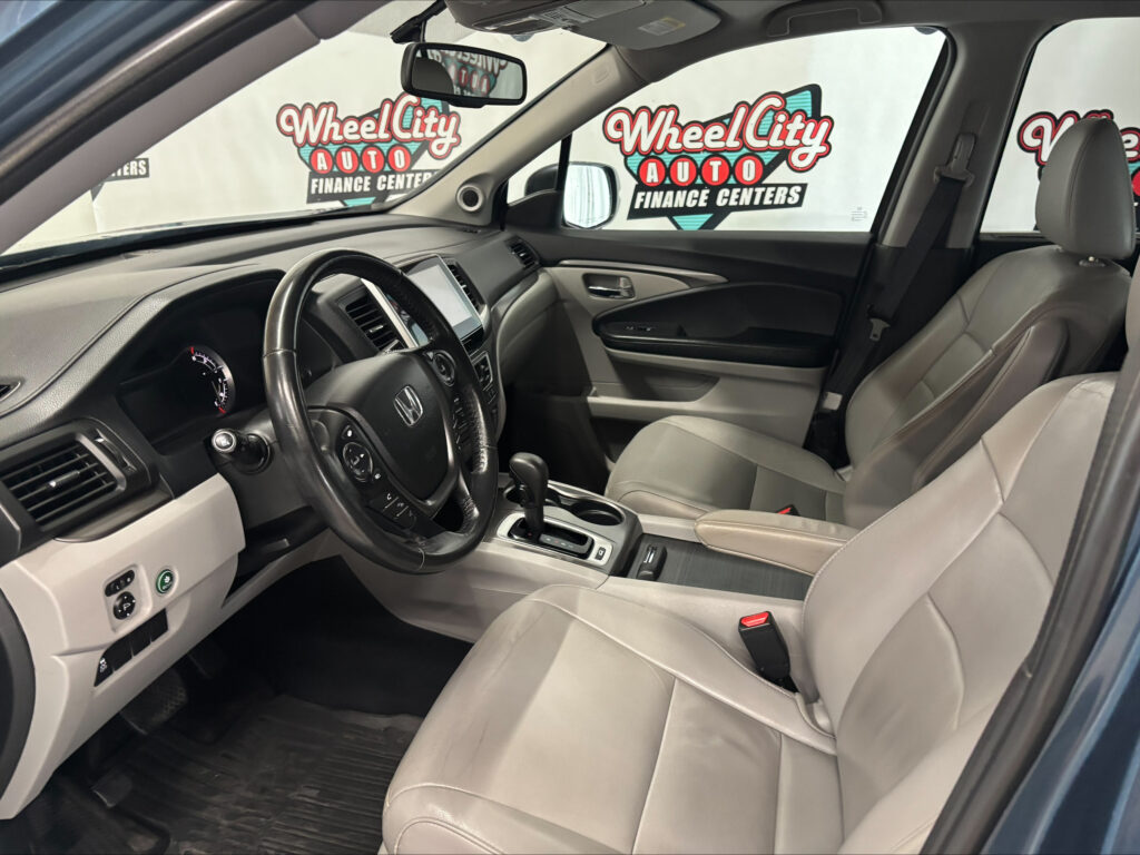 Interior of a gray-tan car showing steering wheel, dashboard, center console, and front/right seats with a dealership backdrop outside the windows