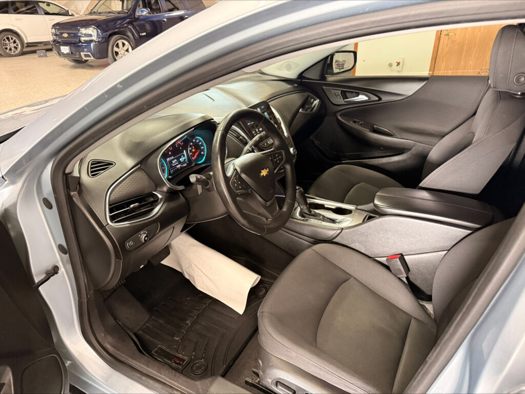 Front interior of a Chevrolet sedan with the driver’s door open, showing the steering wheel, digital gauge cluster, and center console.