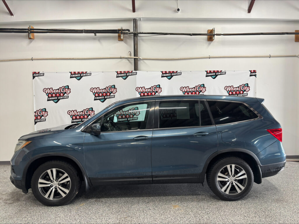 Blue SUV parked indoors in front of a Wheel City Auto Finance Centers banner on a white wall as ceiling pipes loom above it.